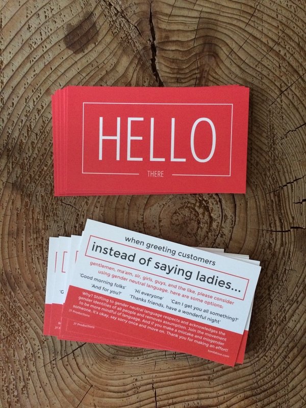 A picture of business cards on a wooden table. The front is red and says Hello There in white text. The back says, instead of using words like ladies and gentleman, guys, sir, ma'am, etc, try using folks, y'all everyone... Using gender neutral language respects and acknowledges a person's gender identity, and removes assumption.