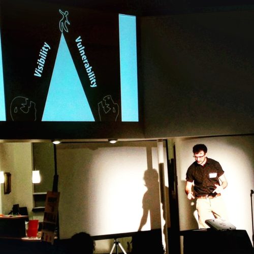 Kingsley is on stage at DisruptHR. Behind him is a slide that says Visibility on one side, and vulnerability on the other. The picture shows a person teetering on top of a triangle (drawn)