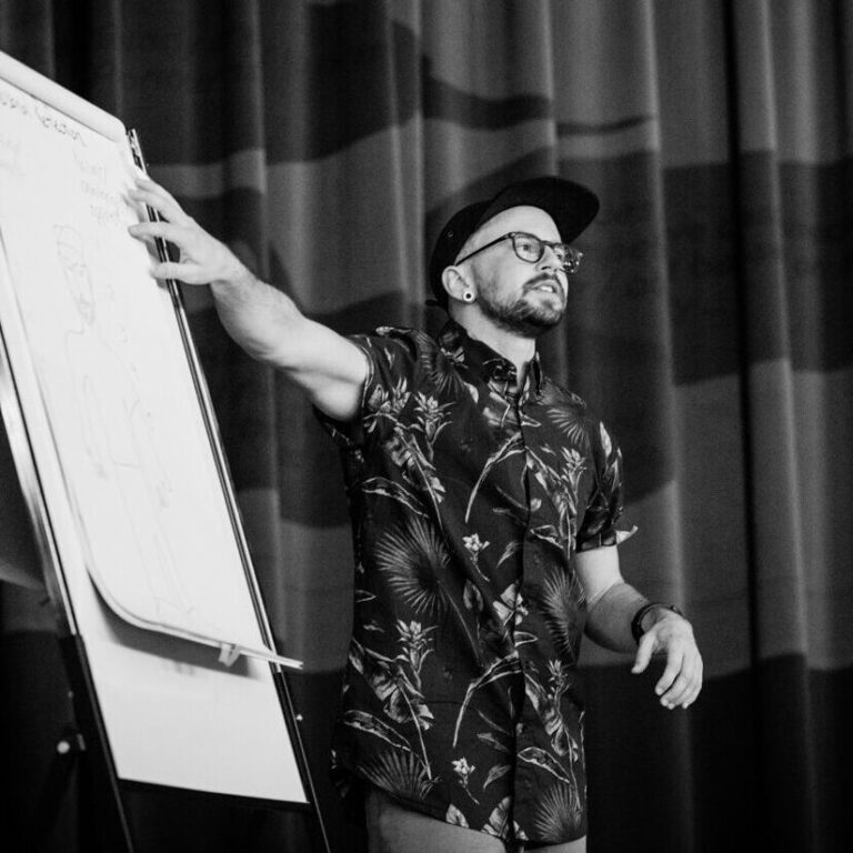 Kingsley stands on a stage with curtains in the background and he gestures to a page of flip-chart paper.
