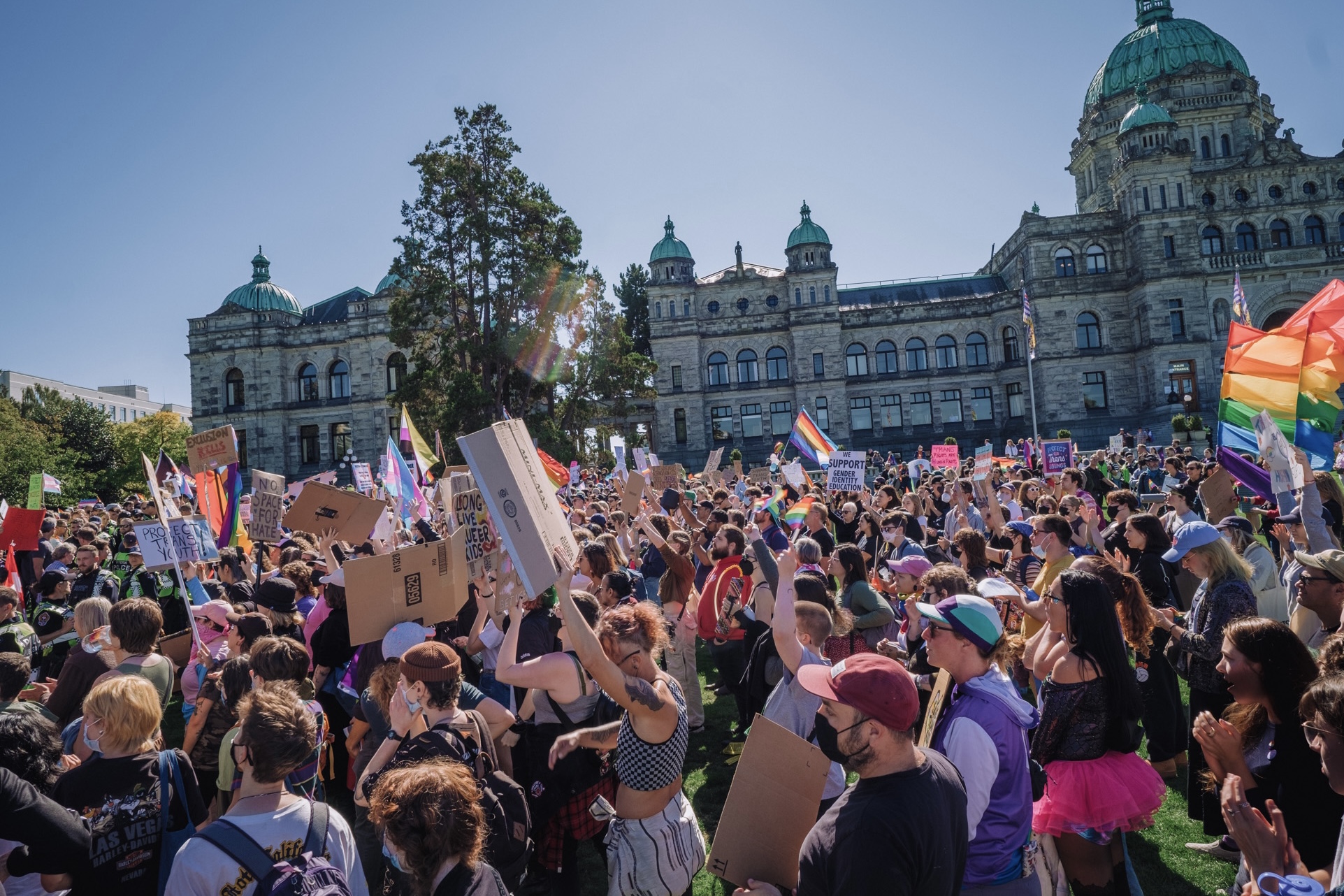 A large crows of counter-protestors holding signs and flags in support of LGBTQ2S+ people. The crowd is standing in front of the British Columbia Legislature.