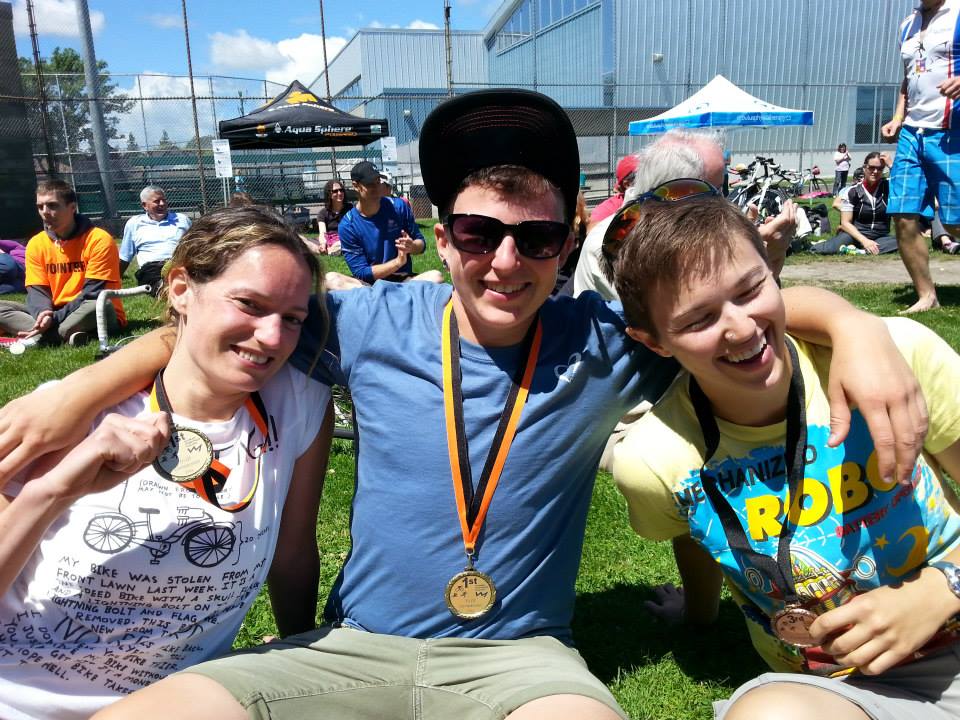 A photo of Kingsley wearing a black ball cap, sunglasses, a blue shirt, and green shorts. Kingsley's arms around the shoulders of two friends, and Kingsley has a medal after completing the Triathlon of Compassion. (2014)